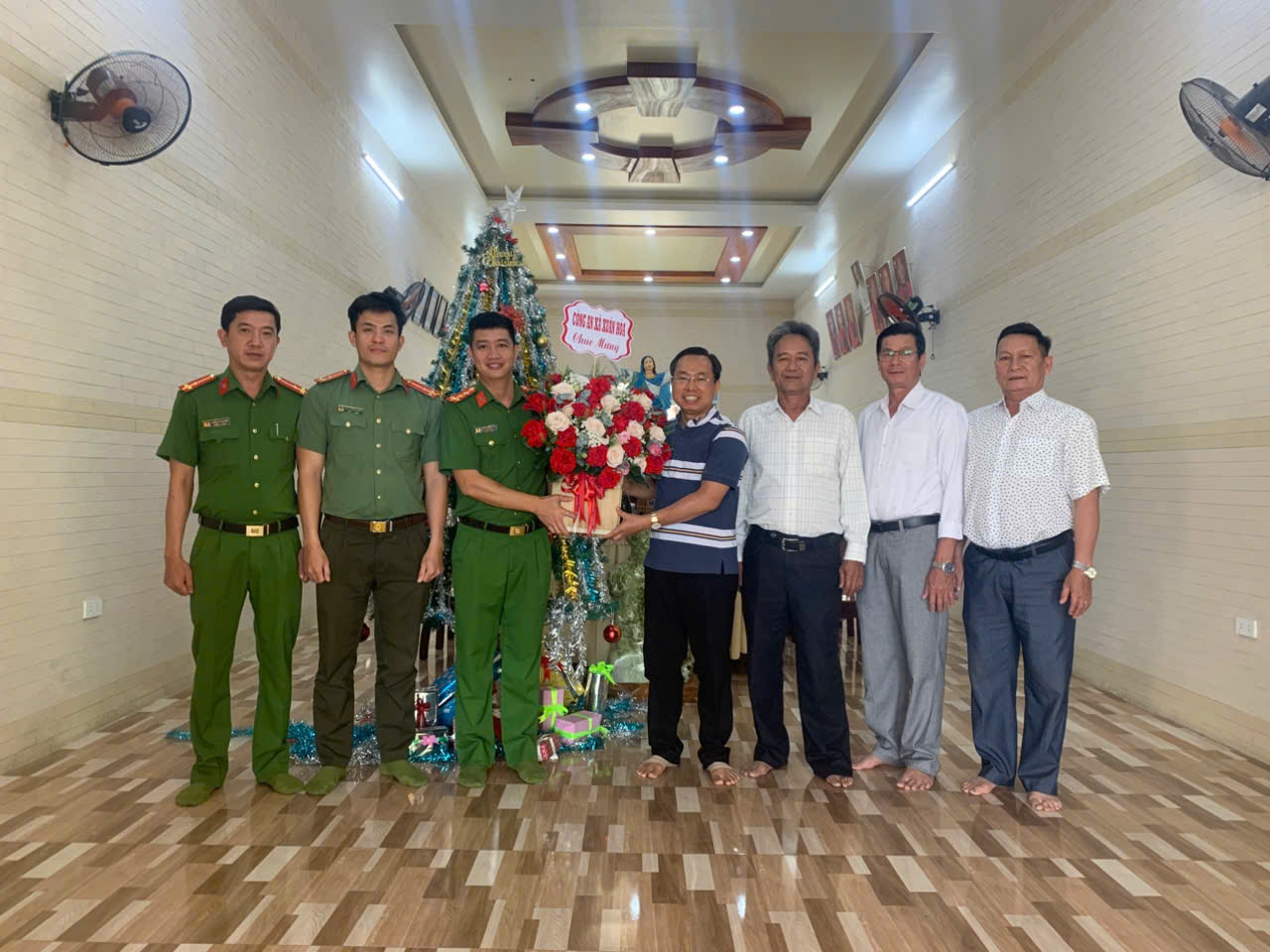 'Description: A group of men standing in a hallway with a christmas tree and a gift

AI-generated content may be incorrect.'
